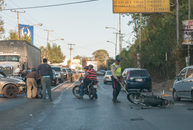 Eine Gruppe von Menschen steht um ein verunglücktes Motorrad auf der Straßenseite mit mehreren Fahrzeugen, darunter ein Lastwagen, und einer Hintergrund von Bäumen, Masten, Lichtern und Schildern unter einem Himmel.