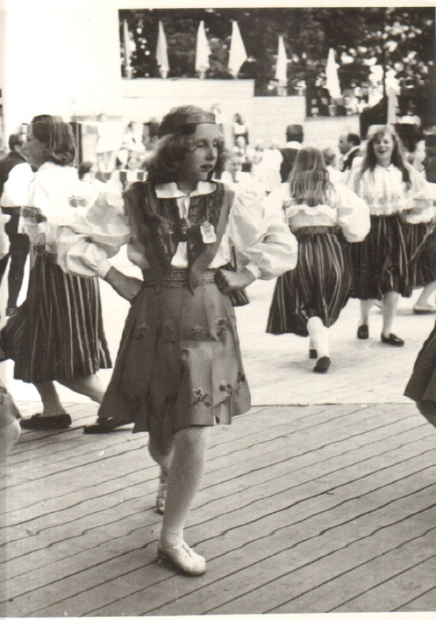Eine Gruppe von Frauen in traditioneller bayrischer Tracht tanzt auf einem Holzboden, mit einer Wand, Bäumen und anderen Gegenständen im Hintergrund. Text ist am unteren Bildrand sichtbar.