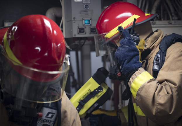 Zwei Feuerwehrleute in Schutzausrüst arbeiten an einem Feuerhydranten während einer Ausbildung mit Maschinen und Kabeln im Hintergrund.