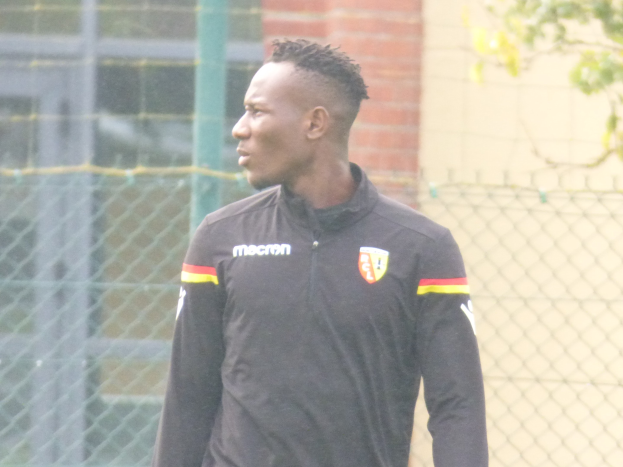 Ein Mann in einer schwarzen Jacke steht auf einem Fussballfeld umgeben von einem Zaun und einem Baum und sieht konzentriert während eines Trainingssitzungs.