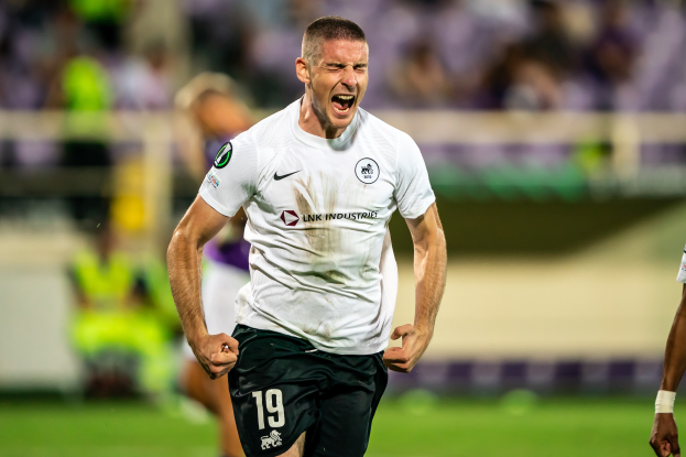 Ein Mann in einem weißen Hemd und schwarzen Shorts rennt auf einem Fußballfeld und feiert mit einer Gruppe von Menschen im Hintergrund.