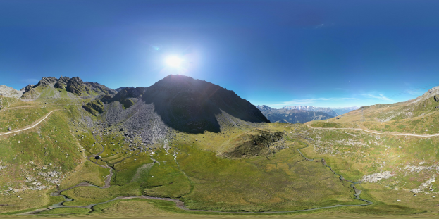 Panoramischer Blick auf eine Bergkette mit grüner Wiese, einer gewundenen Straße und einem hellblauen Himmel unter einer strahlenden Sonne.