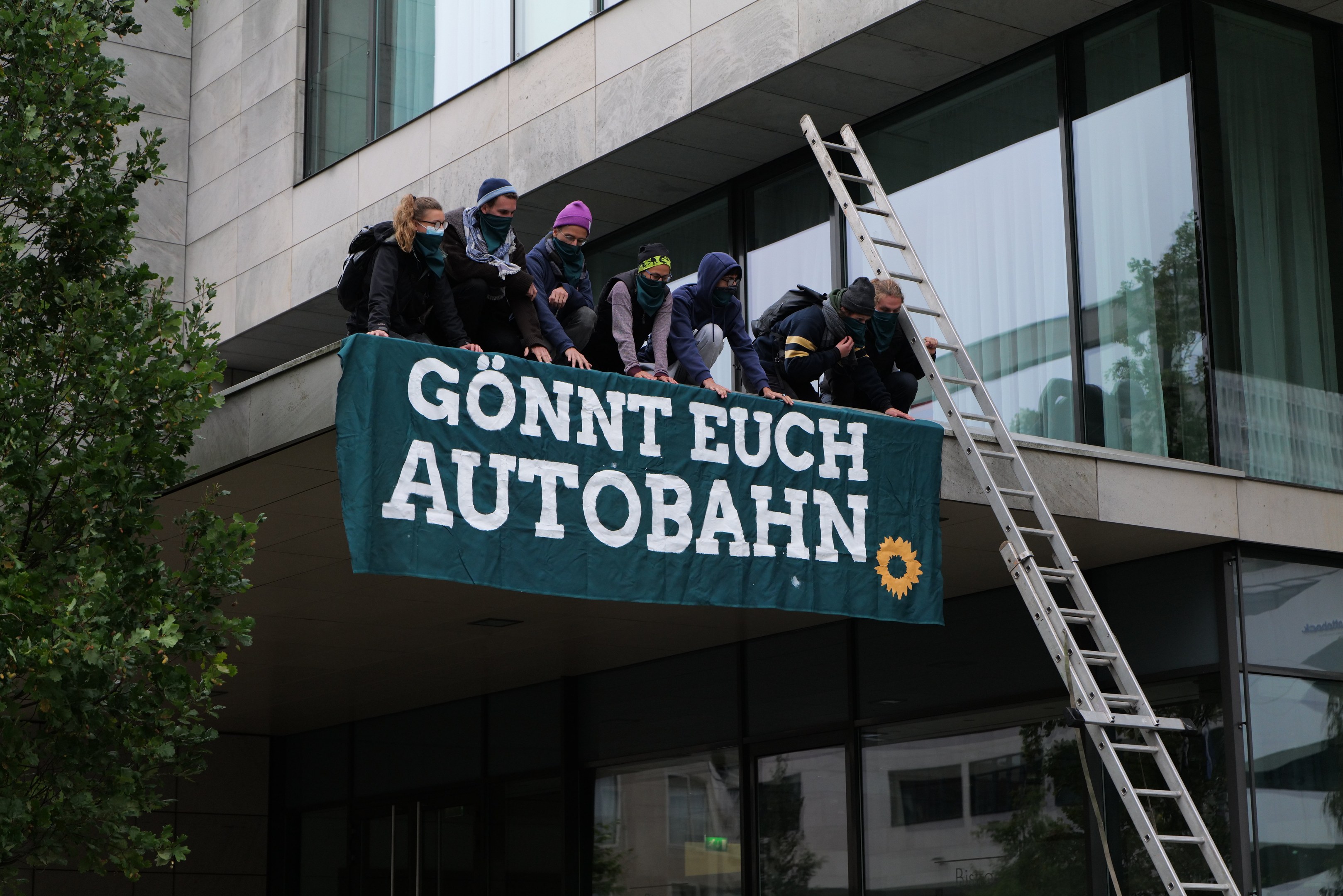 Eine Gruppe von Menschen auf einem Gebäude, das eine Fahne hält, mit einem Baum links und Glasfenstern im Hintergrund, die scheinbar für die deutsche Regierung protestieren.