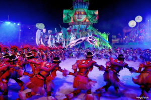 Eine Gruppe von Menschen in bunten Kostümen tanzt auf einer Bühne vor einem sitzenden Publikum, beleuchtet von Scheinwerfern und umgeben von Pfählen und Ballons, mit einem Bildschirm im Hintergrund, der eine Person und andere Gegenstände zeigt, während des Karnevals in Rio de Janeiro 2014.