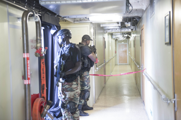 Zwei Polizisten in Uniform stehen nebeneinander in einem Flur, tragen Helme und halten Waffen, mit einer Tür und einem Foto auf der rechten Wand und Deckenlampen und Rohren.