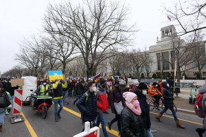Eine große Gruppe von Menschen nimmt an einer Protestdemo auf einer Straße in Washington, D.C. teil, wobei einige Schilder und Banner halten und andere Fahrräder fahren, sowie Schilder, Bäume und einen klaren blauen Himmel im Hintergrund.