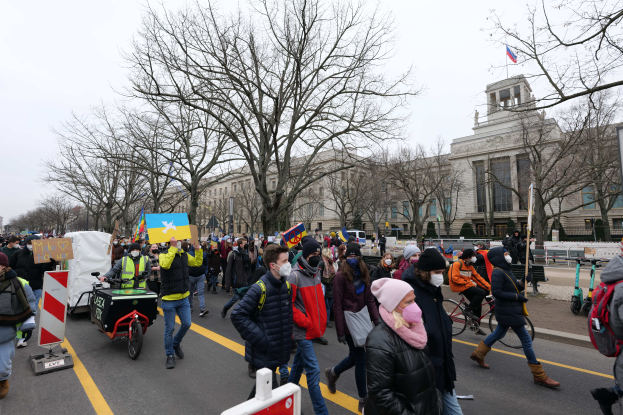 Eine große Gruppe von Menschen nimmt an einer Protestdemo auf einer Straße in Washington, D.C. teil, wobei einige Schilder und Banner halten und andere Fahrräder fahren, sowie Schilder, Bäume und einen klaren blauen Himmel im Hintergrund.