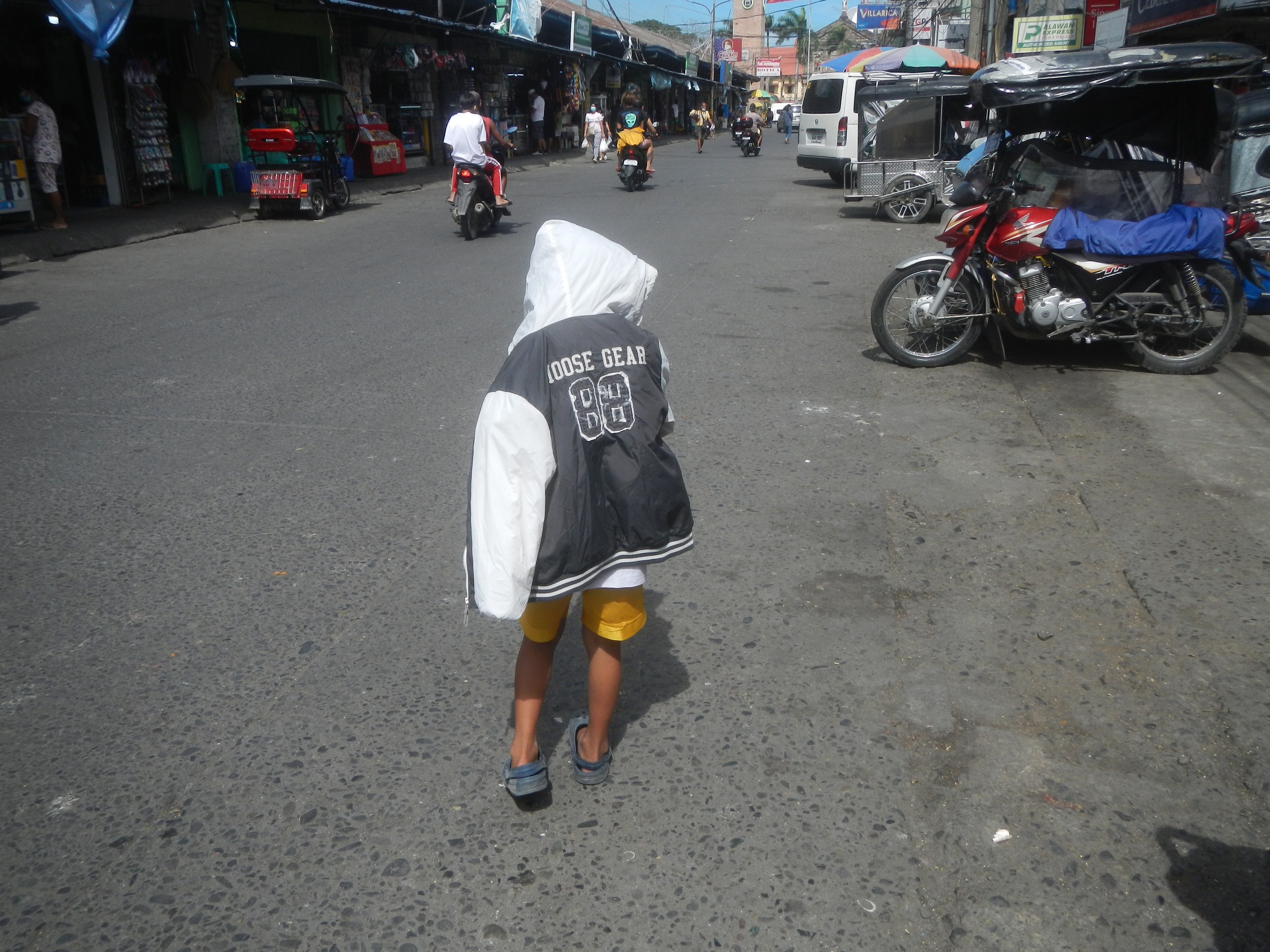 Ein junger Junge geht eine Straße voller Motorräder und anderer Fahrzeuge entlang, mit Gebäuden, Bäumen und einem klaren Himmel im Hintergrund.