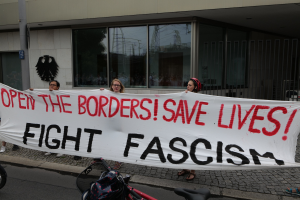 Eine Gruppe von Menschen mit einem Transparent mit der Aufschrift 'öffne die Grenzen, rette Leben, bekämpfe Faschismus' vor einem Gläserfenster und einer Stange, mit einem Fahrrad und einer Tasche auf der Straße.