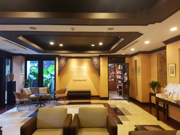 Ein geräumiger Hotelfoyer mit Sitzbereichen, Dekorationspflanzen, Bücherregalen, Wandkunst und Seitentisch-Dekoration, beleuchtet von Deckenlampen und natürlichem Sonnenlicht durch Fenster und Türen.