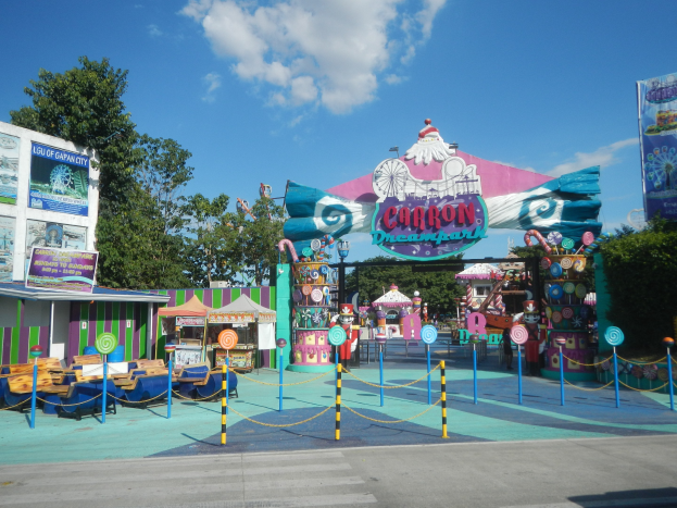 Achterbahn am Eingang eines Parks mit Straße, eingezäuntem Bereich, Buden, Banner, Bäumen und bewölktem Himmel im Hintergrund.