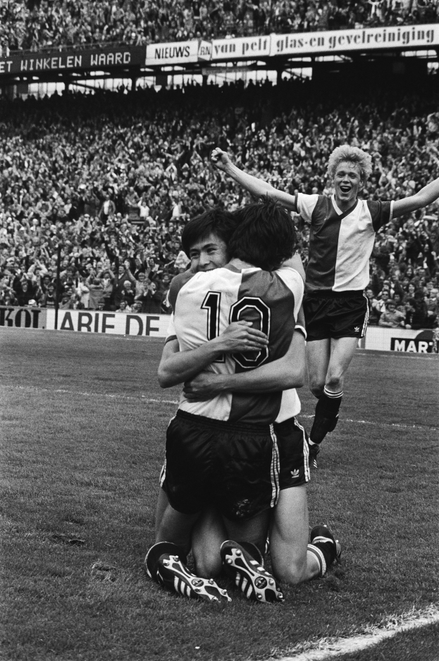 Drei Fußballspieler feiern auf dem Boden nach einem Tor, mit jubelnder Menge und schwenkenden Fahnen im Hintergrund, in Schwarz-Weiß aufgenommen.