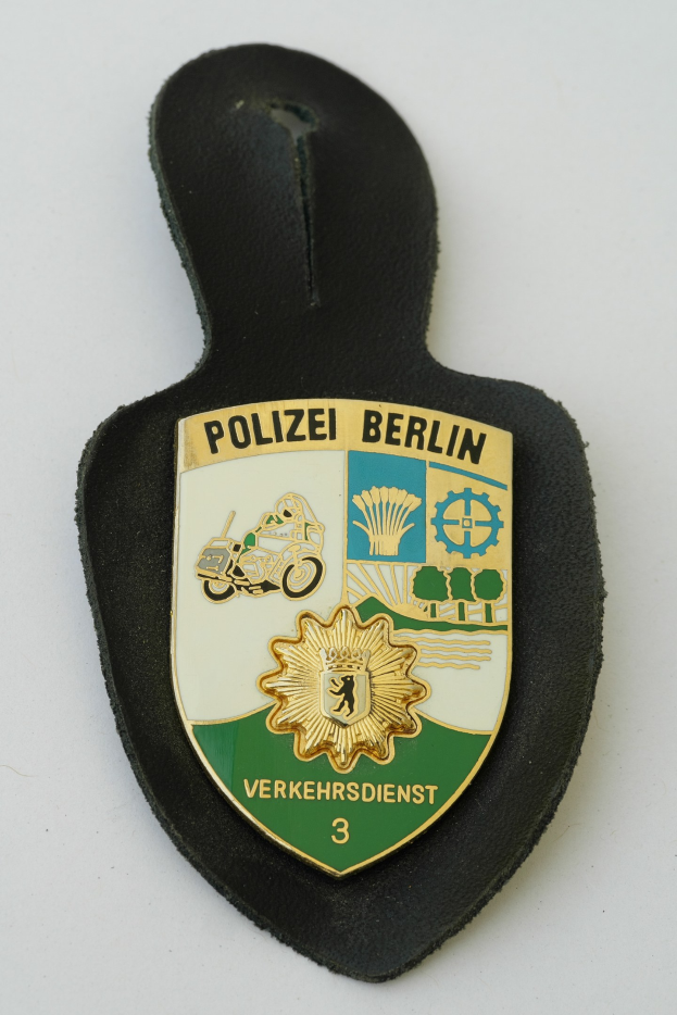 Ein schildförmiges Polize abadge mit 'Polizei Berlin' in schwarzer Schrift auf einem blauen Hintergrund, das ein weißes Stern und einen weißen Rand hat, auf einer Oberfläche platziert.