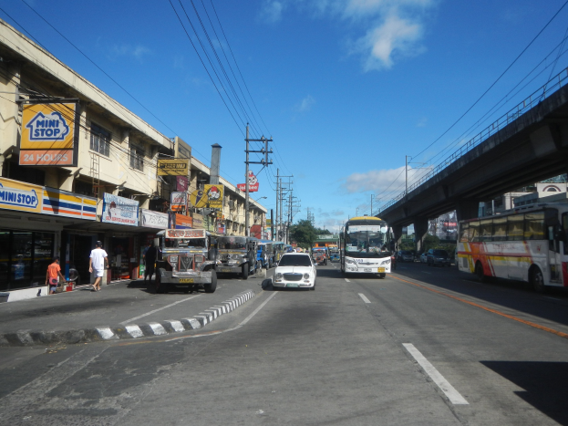 Eine belebte Stadtstraße mit Autos, Bussen, Fußgängern auf dem Gehweg, Gebäuden, Strommasten, einer Brücke, Bäumen und einem bewölkten Himmel.