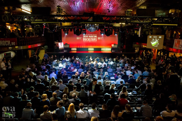 Eine große Menschenmenge sitzt auf Stühlen vor einer Bühne mit Bildschirmen und Bannern, beleuchtet von Scheinwerfern und umgeben von Geländern, mit einem Wasserzeichen in der linken unteren Ecke, in Erwartung des Beginns eines Online-Marketing-Rockstars-Events.