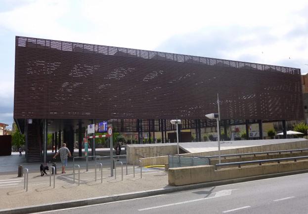 Großes modernes Gebäude mit zahlreichen Fenstern, das als neuer Eingang zur Stadt dient, flankiert von Straßeninfrastruktur, Fahrzeugen, Fußgängern und Bäumen unter einem bewölkten Himmel.