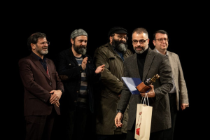 Eine Gruppe von Mönnen steht auf einer Bühne, einer hölt eine Tasche und eine Trophäe, mit einem dunklen Hintergrund, der die Eröffnung der Berliner Filmfestspiele suggeriert.