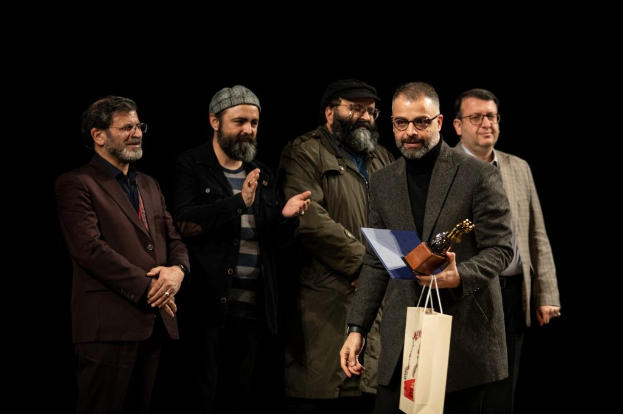 Eine Gruppe von Mönnen steht auf einer Bühne, einer hölt eine Tasche und eine Trophäe, mit einem dunklen Hintergrund, der die Eröffnung der Berliner Filmfestspiele suggeriert.