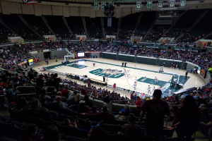 Eine große Arena mit Zuschauern, die ein Basketballspiel beobachten, mit einem Spielfeld in der Mitte und Anzeigetafeln, Lampen und anderen Objekten oben.