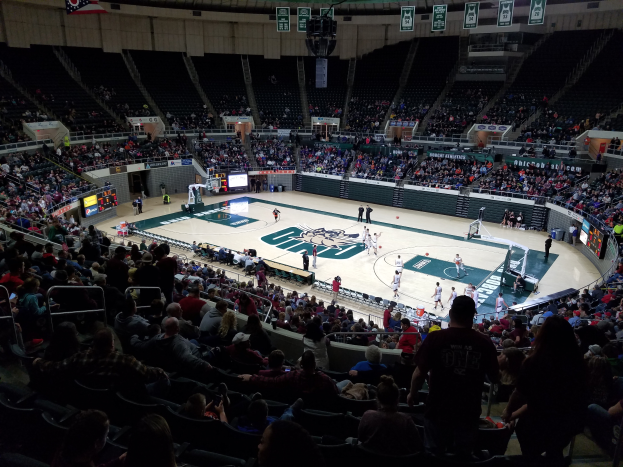 Eine große Arena mit Zuschauern, die ein Basketballspiel beobachten, mit einem Spielfeld in der Mitte und Anzeigetafeln, Lampen und anderen Objekten oben.