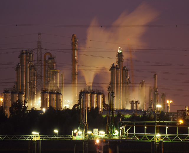 Ölraffinerie bei Nacht mit Rauchentwicklung, beleuchtet von nahen Lichtern, mit sichtbarem Himmelhintergrund und Wasserzeichen.