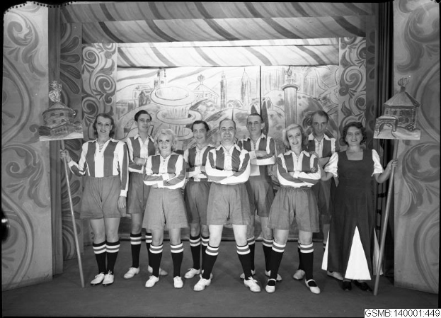 Eine Gruppe strahlender Frauen in 40er-Jahre-Kleidung steht auf einer Bühne und hält Stöcke in den Händen, mit einem Gemälde an der Wand dahinter und einem Text am unteren Rand, der '40er-Jahre-Frauenfußballmannschaft' lautet.