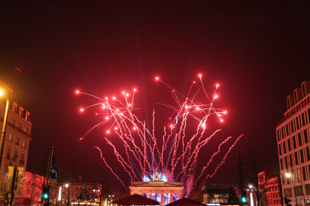 Eine Stadtstraße in Berlin an Silvester, gefüllt mit Gebäuden, Bäumen, Laternenpfählen, Verkehrszeichen, Zeltplanen und Menschen, mit einem von Feuerwerk erleuchteten Himmel im Hintergrund.