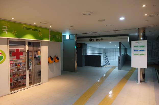 Apothekeneingang mit einem "Apotheke"-Schild auf der linken Seite, einer Tafel mit Text auf der rechten Seite, beleuchteten Lampen oben und Hintergrundobjekten.