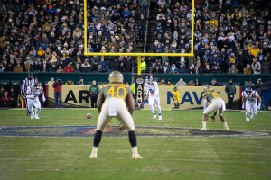 Fußballspieler in Sportkleidung und Helmen konkurrieren auf einem Feld mit Zuschauern im Hintergrund, Pfosten und Tafeln mit Text.