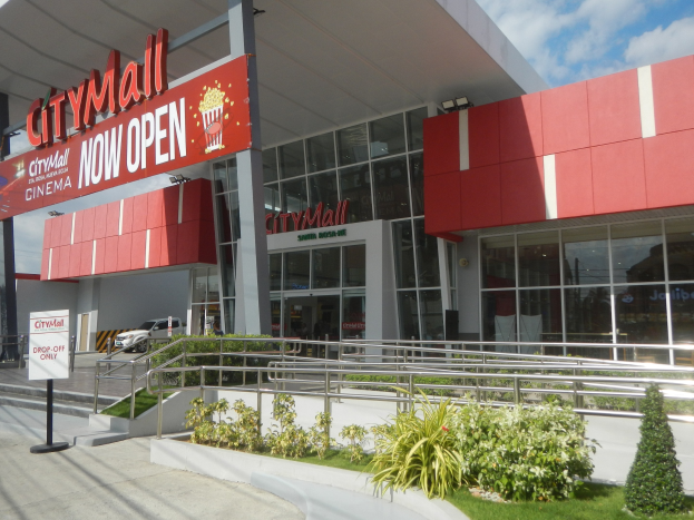 Stadtzentrum-Eingangsschild mit Aufschrift "Stadtzentrum jetzt geöffnet" mit umgebender Vegetation, Metallzaun, Menschen, Auto, Pfosten, Banner, Lichter und bewölktem Himmel.