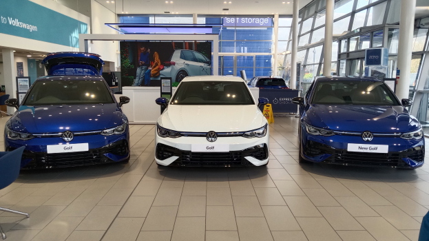 Volkswagen-Ausstellung mit drei blauen und weißen Autos, einem Tisch mit Gegenständen, Informationsplakaten, Glaswänden und Deckenleuchten.