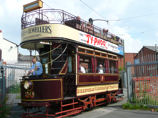 Ein Doppeldeckerbus mit Menschen an Bord fährt eine Straße entlang, umgeben von einem Metallzaun, Pflanzen mit Blumen, einem Schild, einem Straßenpfosten, einigen Drähten, einer Gruppe von Gebäuden mit Fenstern und einem bewölkten Himmel.