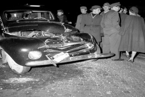 Eine schwarz-weiße Aufnahme eines beschädigten Autos am Straßenrand, umgeben von einer Gruppe besorgter Menschen.