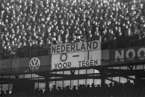 Ein Schwarz-Weiß-Foto einer großen Menschenmenge in einem Stadion mit einer Texttafel in der Mitte und ein paar Individuen am Boden.