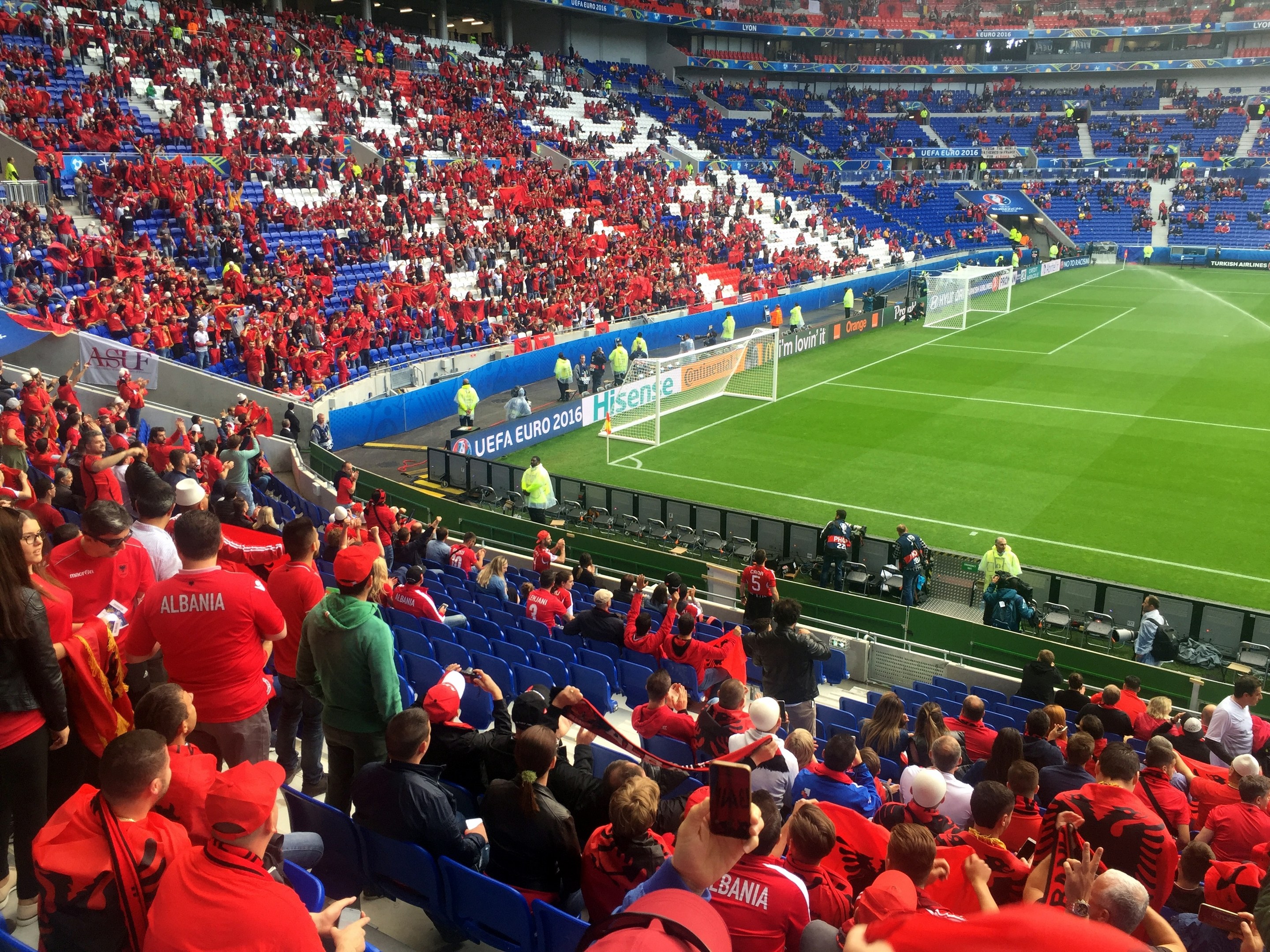 Eine große Menschenmenge in einem Stadion schaut einem Fußballspiel zu, mit Menschen auf Stühlen und Stehplätzen, Fahnen, einem Torpfosten und Deckenbeleuchtung.