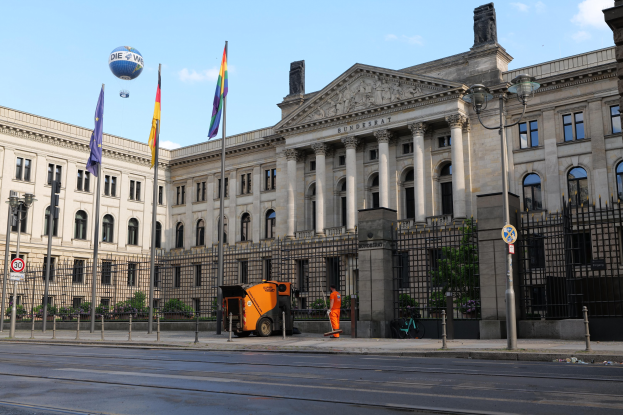 Großes modernes Gebäude mit vielen Fenstern, als Bundestag in Berlin identifiziert, umgeben von Straßeninfrastruktur, Fußgängern, Flaggen und Bäumen unter einem bewölkten Himmel.