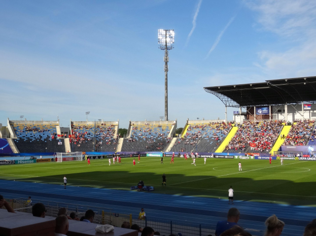 Ein Fußballspiel in einem großen, flutlichtbeleuchteten Stadion mit Zuschauern auf den Rängen und Spielern auf dem Feld unter einem sichtbaren Himmel.