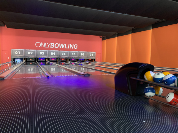 Eine Bowlingbahn mit einer Reihe von Bowlingkugeln auf der rechten Seite, aufgestellten Kegeln in der Mitte und einer textbedeckten Wand im Hintergrund, mit der Decke darüber.