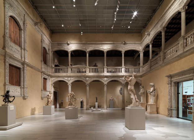 Ein geräumiger Museumsausstellungsraum mit zahlreichen Statuen und Skulpturen auf Sockeln, architektonischen Elementen wie Wänden, Säulen, Geländern und Deckenleuchten sowie einigen Besuchern, die die Kunstwerke betrachten.