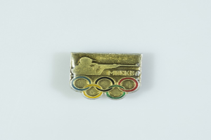 Eine goldene und silberne olympische Abzeichen-Nadel mit den olympischen Ringen und dem Text 'Olympische Spiele' darauf, vor einem weißen Hintergrund.