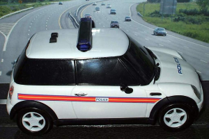 Weißer Polizeiwagen mit "Polizei"-Aufschrift auf einer Straße außerhalb der Stadt, umgeben von fließendem Verkehr, Gras und Bäumen auf beiden Seiten.