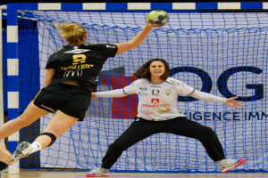 Zwei Frauen beim Handballspiel vor einem Tor, eine hält den Ball, mit Netz und einer Texttafel im Hintergrund.