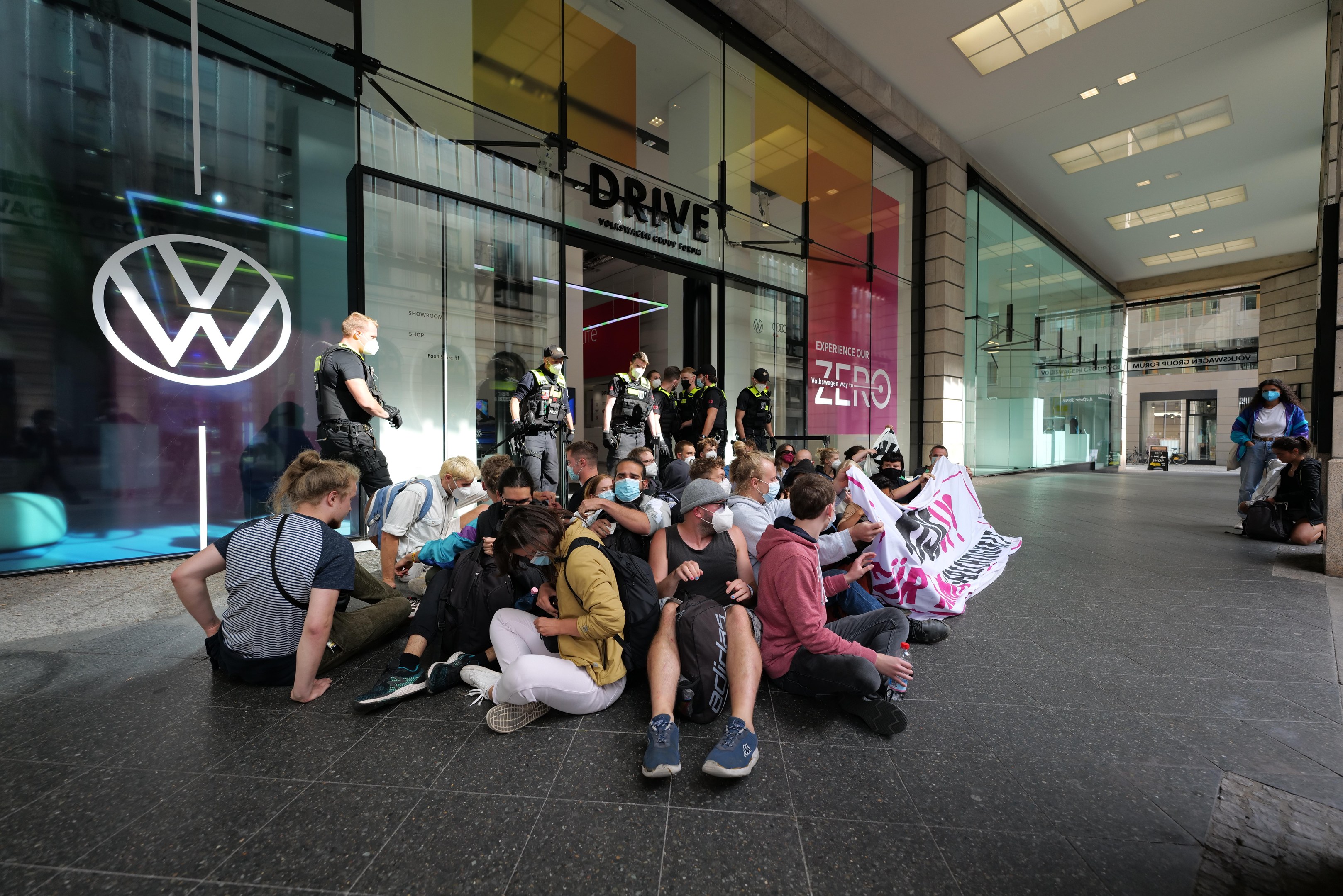 Gruppe von Menschen, die vor einer Volkswagen-Niederlassung auf dem Boden sitzen und eine Fahne halten.