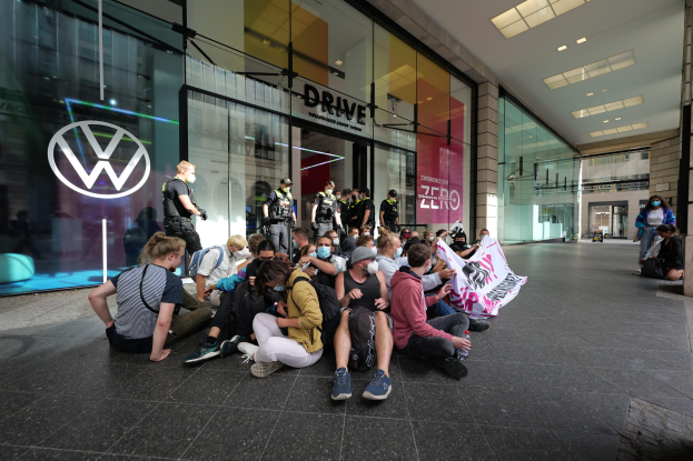 Gruppe von Menschen, die vor einer Volkswagen-Niederlassung auf dem Boden sitzen und eine Fahne halten.