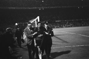 Eine Gruppe von Männern, die auf einem Fußballfeld mit einem Torpfosten und beleuchteten Tribünen im Hintergrund feiern, in Schwarz-Weiß aufgenommen.