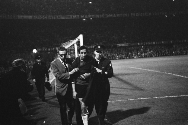 Eine Gruppe von Männern, die auf einem Fußballfeld mit einem Torpfosten und beleuchteten Tribünen im Hintergrund feiern, in Schwarz-Weiß aufgenommen.