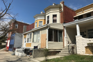Eine Reihe von Häusern in einem schlechten Zustand, einige vernagelt, mit Glasfenstern, Treppen mit Geländern, Pflanzen, Gras, Bäumen und einem bewölkten Himmel im Hintergrund.