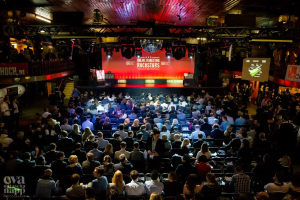 Eine große Menschenmenge sitzt auf Stühlen vor einer Bühne mit Bildschirmen und Bannern, beleuchtet von Scheinwerfern und umgeben von Geländern, mit einem Wasserzeichen in der linken unteren Ecke, in Erwartung des Beginns eines Online-Marketing-Rockstars-Events.