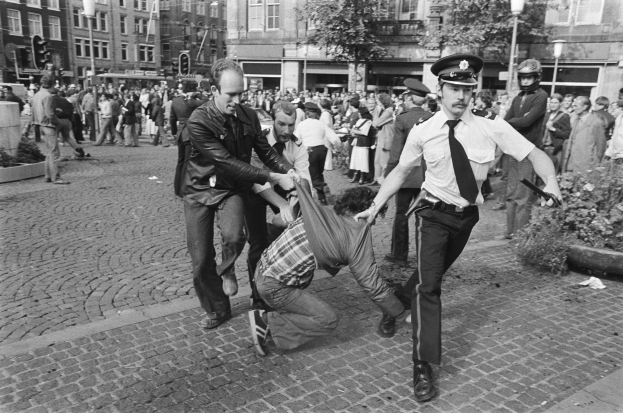 Schwarz-weißes Bild eines Mannes, der von zwei Polizisten auf einer Straße gezogen wird, mit Zuschauern, Bäumen, Gebäuden, Laternen, Verkehrsampeln, Topfpflanzen und einem Mann in Schuhen im Vordergrund.
