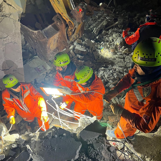 Eine Gruppe von Menschen in orangefarbenen Uniformen und grünen Helmen bei der Arbeit an Schutt von einem eingestürzten Gebäude, mit Werkzeugen und Materialien, und einem Kran im Hintergrund.